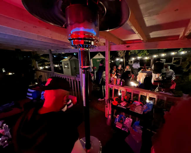 A gathering on a wooden patio, with people seated around tables and a patio heater illuminating the scene under string lights.