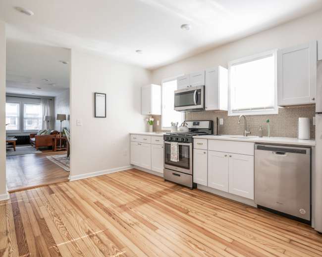 The image shows a modern kitchen with wooden floors, white cabinetry, stainless steel appliances, and a window allowing natural light.