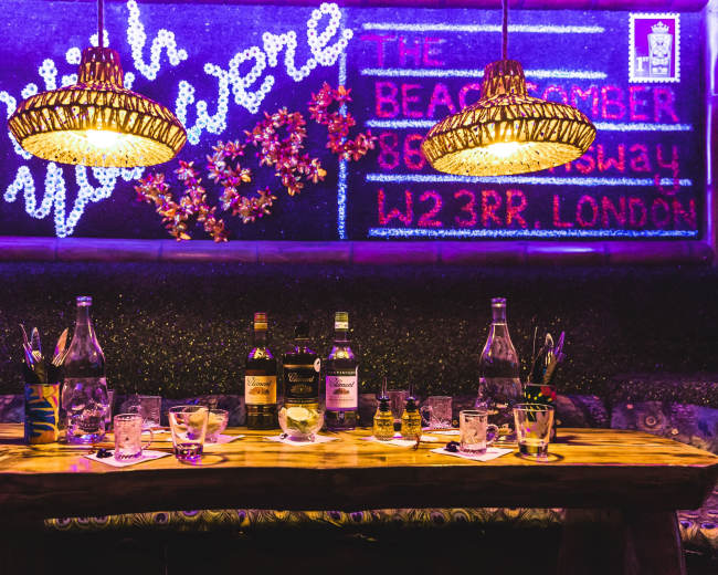 A wooden table is set with various bottles of alcohol and glasses, under two decorative lamps in a dimly lit bar adorned with colorful signage.