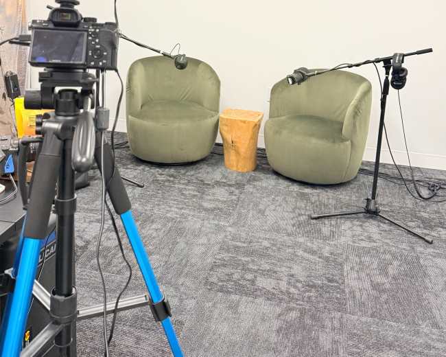 The image shows a podcast recording setup featuring two green chairs, a small wooden table, and a camera on a tripod pointing towards the chairs.