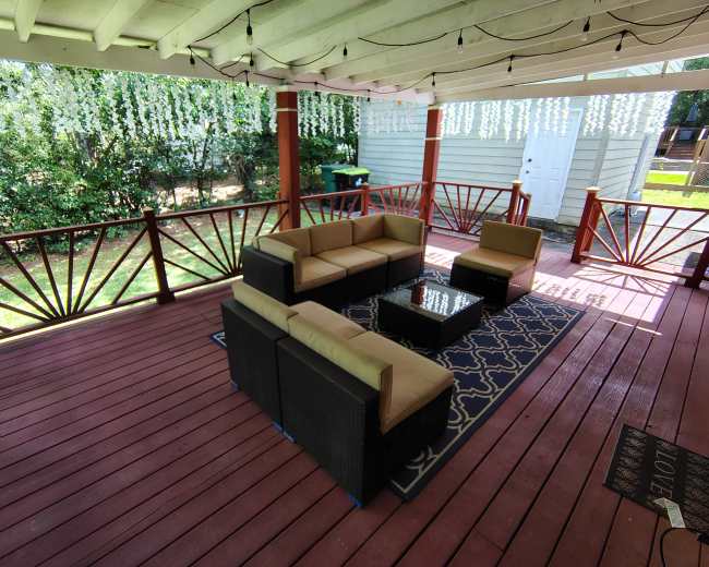 A covered patio features a seating area composed of a sectional sofa and two chairs, arranged around a coffee table on a patterned rug.