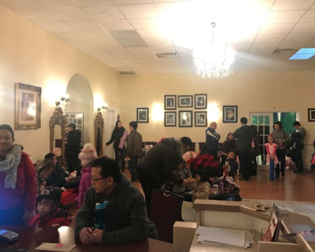 A group of people, including children, are gathered in a spacious room adorned with framed photographs and a chandelier, while some are seated and others are standing.