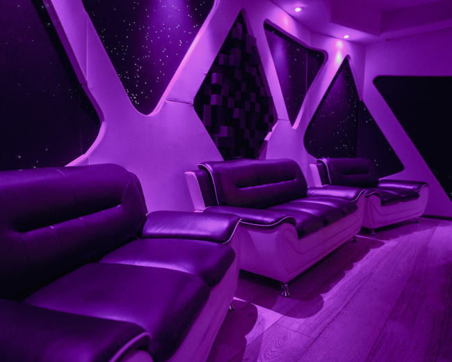 The image shows a modern interior with black and white sofas against a dark, starry wall, illuminated by purple lighting.