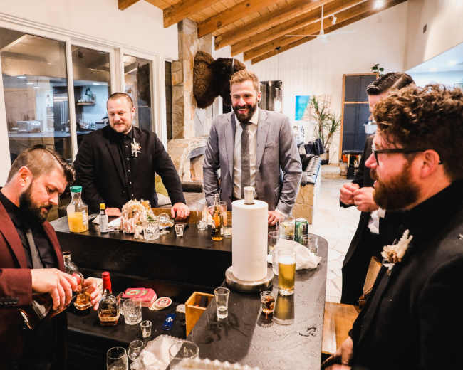 A group of well-dressed men gathers around a bar, with one man pouring a drink while others engage in conversation.