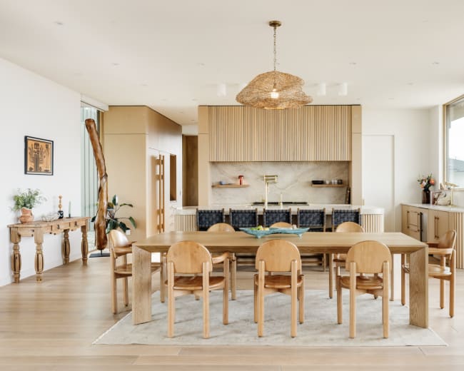The image shows a modern dining area featuring a long wooden table surrounded by eight round chairs, with a large window and an open kitchen in the background.
