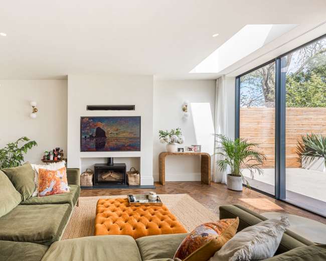A modern living room features a green sectional sofa, an orange ottoman, a wall-mounted television, and large windows opening to a patio.