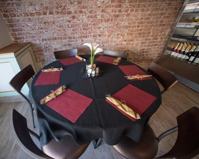 A round table is set with maroon placemats and utensils, surrounded by wooden chairs in a restaurant with a brick wall.