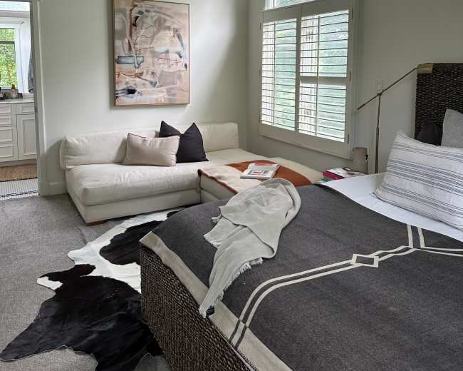 A modern bedroom features a bed with a dark throw blanket, a leather couch, and a large window with shutters, showcasing a minimalist design.