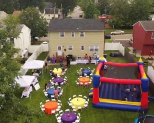The scene shows a backyard party with colorful round tables, chairs, a bounce house, and catering tents set up for guests.