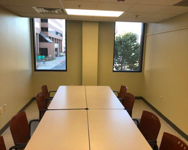 A meeting room with a long table surrounded by several chairs and large windows on two walls.