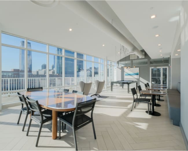 The image shows a well-lit modern lounge area with large windows, featuring a dining table, chairs, and a pool table, overlooking a city skyline.