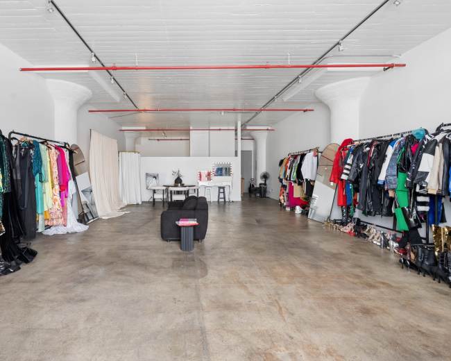 The image shows a spacious, well-lit clothing store or studio with racks of various garments on display along the walls and a table set in the center of the room.