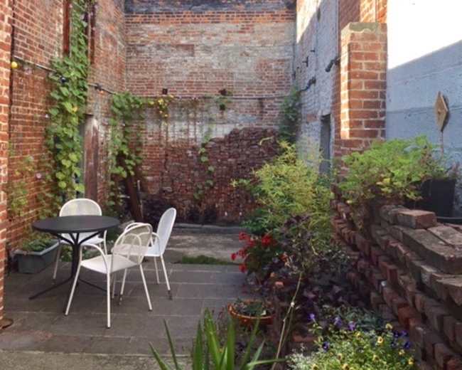 The image shows a small, brick-walled courtyard filled with greenery and a table set with white chairs.
