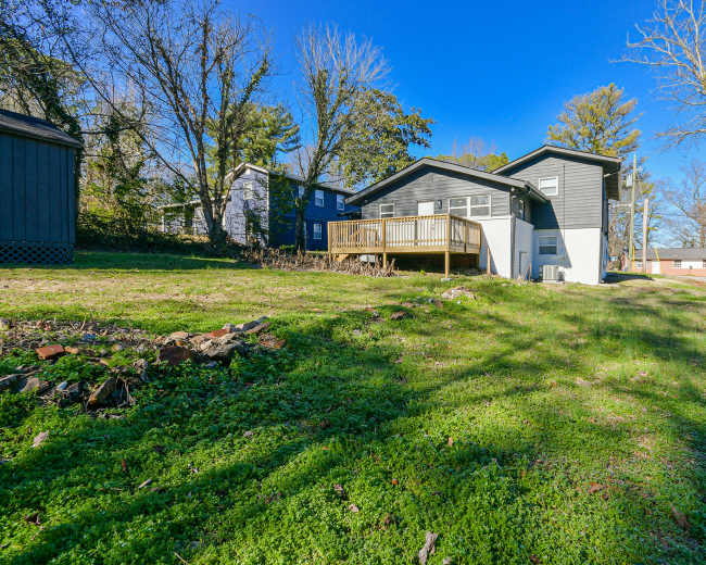 A dark-colored house with a wooden deck is situated in a grassy yard surrounded by trees and other buildings.