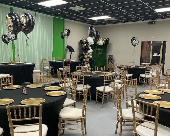 The room is set up for an event with black and gold themed tables and decorations, featuring round tables with black tablecloths and gold plates, surrounded by gold chiavari chairs.