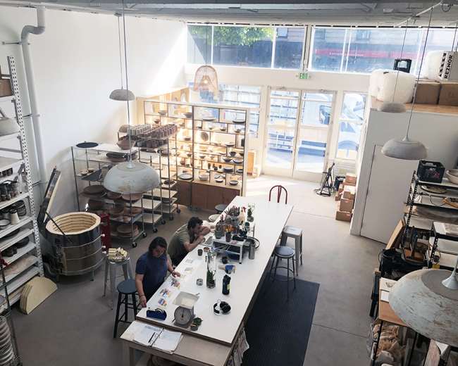 The image shows a pottery studio with two people working at a central table surrounded by shelves filled with ceramic pieces and tools.