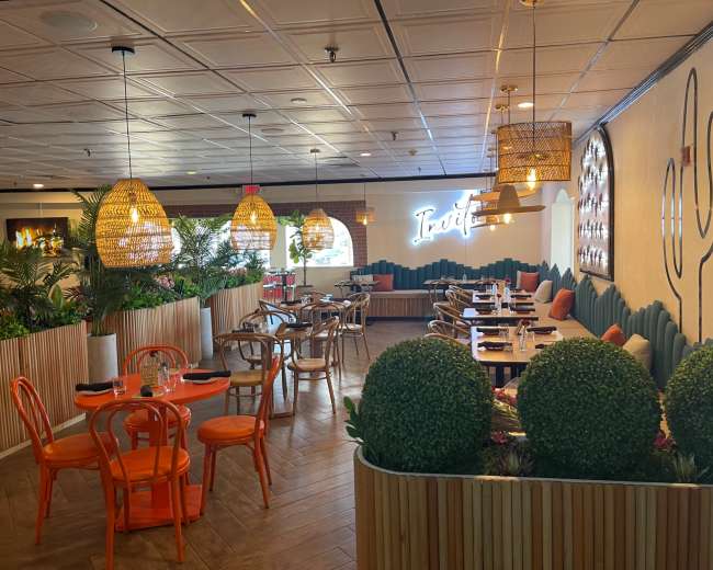 The image shows a modern restaurant interior featuring wooden furnishings, vibrant orange chairs, and greenery decor.
