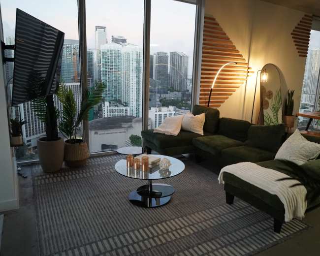 A modern living room features a green sectional sofa, a round glass coffee table with candles, and large windows showcasing a city skyline.