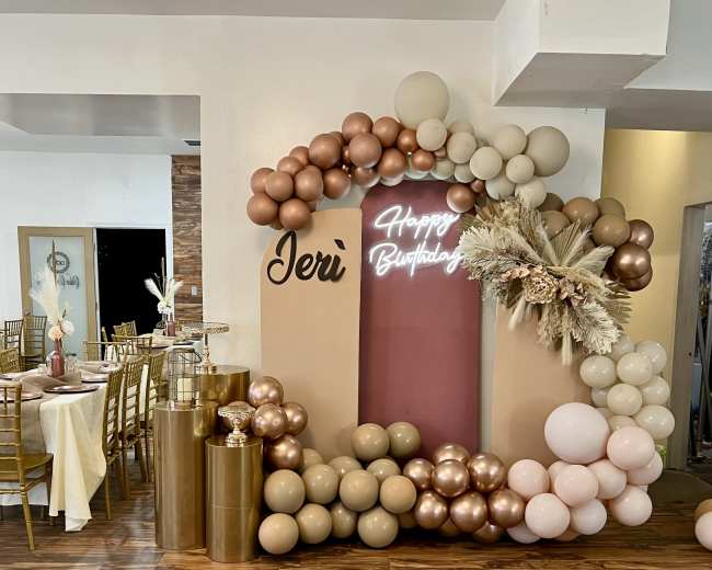 The image shows a festive birthday decoration featuring an arch made of balloons in shades of beige and pink, with a neon sign that reads "Happy Birthday" and elegant table settings in the background.