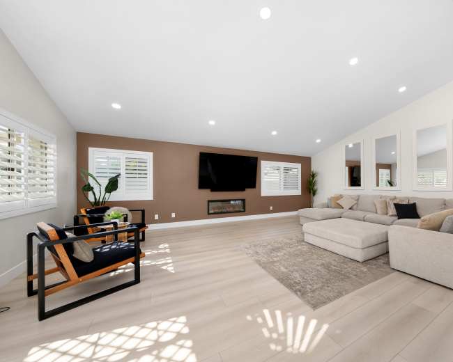 The image shows a modern living room with a sectional sofa, chairs, a television mounted on the wall, and large windows allowing natural light to enter.