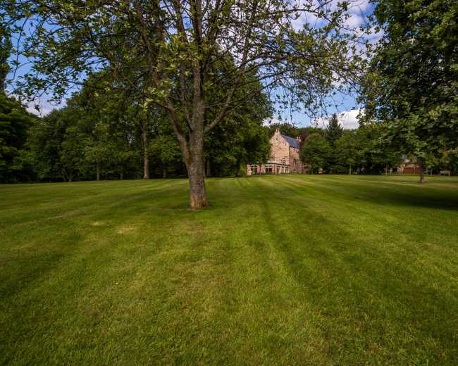 A large, open lawn with manicured grass leads to a stone house partially obscured by trees.