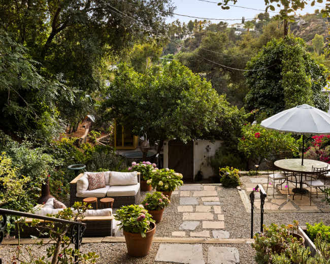 The image shows a landscaped garden with stone pathways, potted plants, a seating area with a sofa, a round dining table under an umbrella, and surrounding greenery.