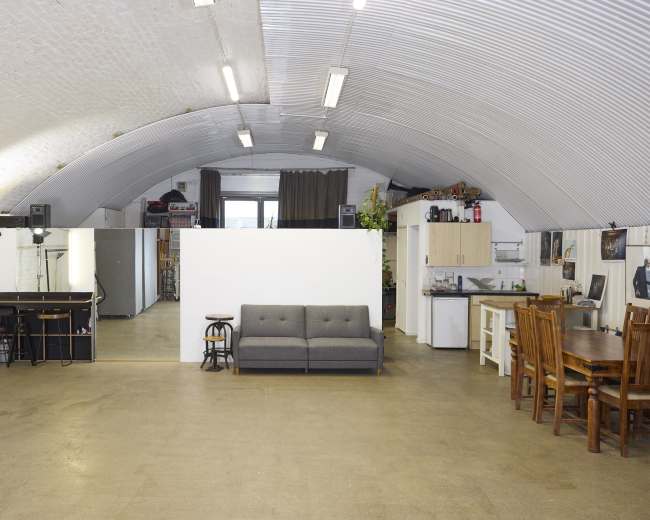 The image shows a spacious, industrial-style loft with an arched ceiling, featuring a living area with a couch and dining space, separated by a white partition.