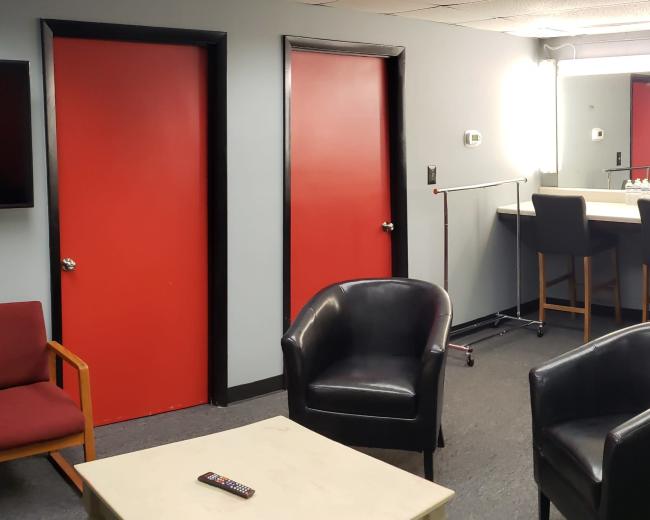 A lounge area with two red doors, a black sofa, several black chairs, a bar-style counter with high stools, and a television mounted on the wall.