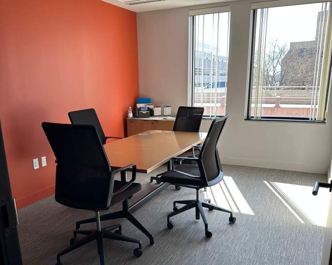 The image shows a bright, empty conference room with a wooden table and four black chairs, set against an orange accent wall and large windows letting in natural light.