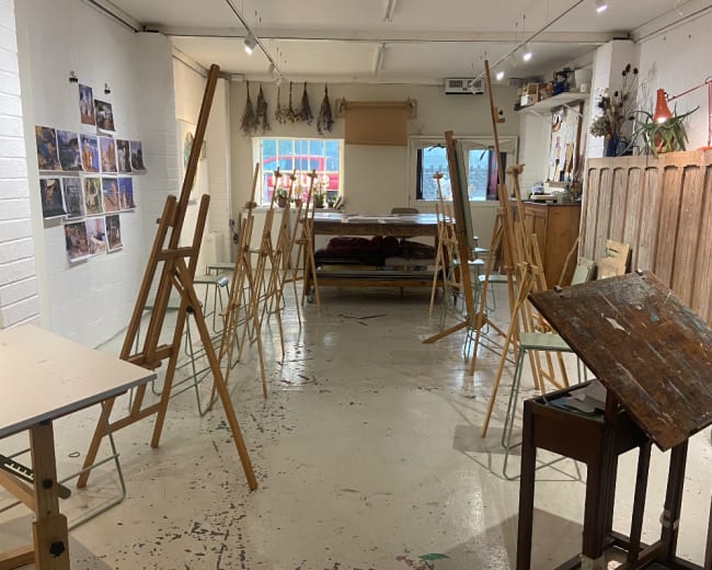 The image shows an art studio with several wooden easels arranged throughout the space, along with a wooden table and various artworks displayed on the walls.