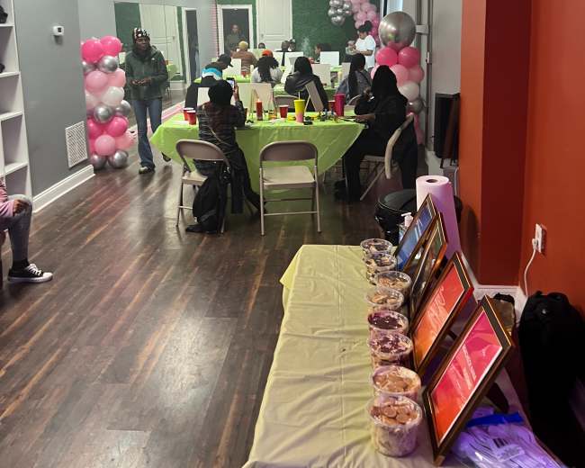 A decorated event space with tables covered in bright green cloth, a buffet of snacks, and balloons along the walls, while guests sit around enjoying the gathering.