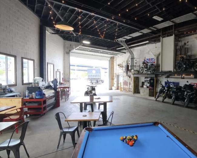The image shows a spacious garage-like interior with a pool table in the foreground, motorcycle displays along the walls, and large windows opening to an outdoor area.