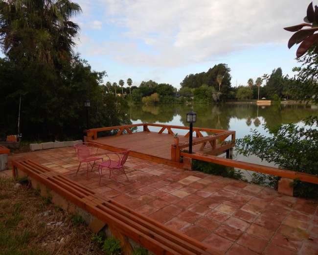 A wooden deck with pink chairs overlooks a calm lake surrounded by greenery and palm trees.