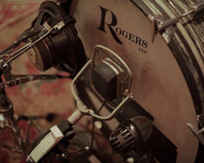 The image features a close-up of a drum kit with various microphones positioned near the bass drum.