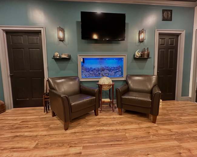 A waiting area with two dark leather chairs, a small round table between them, a large wall-mounted television, and a window displaying an aquarium, all set against teal and dark walls.