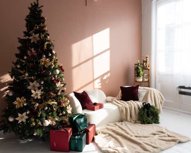 A decorated Christmas tree stands next to a white armchair draped with a blanket, surrounded by wrapped gifts in a softly lit room.