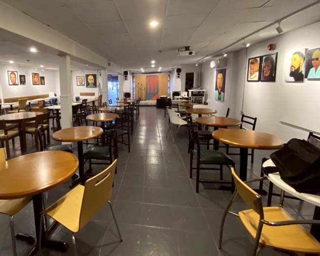 The image shows a spacious indoor café or event space with tables and chairs arranged around the room, featuring artwork on the walls.