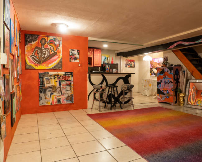 The image shows a colorful basement living space with a gradient rug, a kitchenette in the background, and walls adorned with various pieces of abstract artwork.