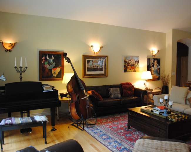 A living room with a piano, a double bass, two sofas, several framed artworks on the walls, and a wooden coffee table in the center.