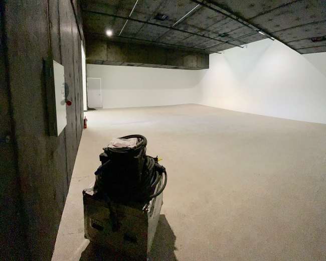 The image shows a large, unfinished room with bare concrete walls and a flat, empty floor.