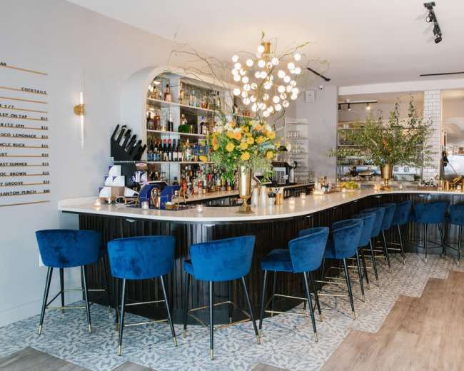 The image depicts a stylish bar area featuring a curved counter with blue upholstered stools, a well-stocked shelf of spirits, and decorative floral arrangements.