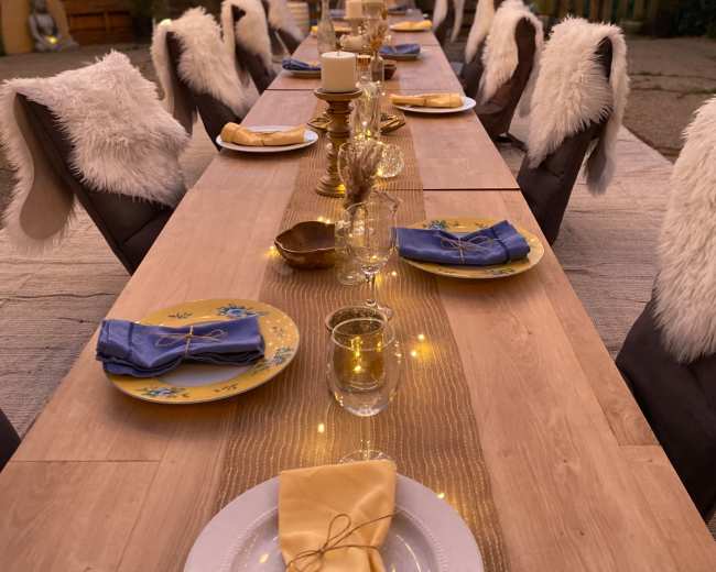 A long outdoor dining table is elegantly set with plates, glasses, and folded napkins, surrounded by chairs adorned with white fur throws.