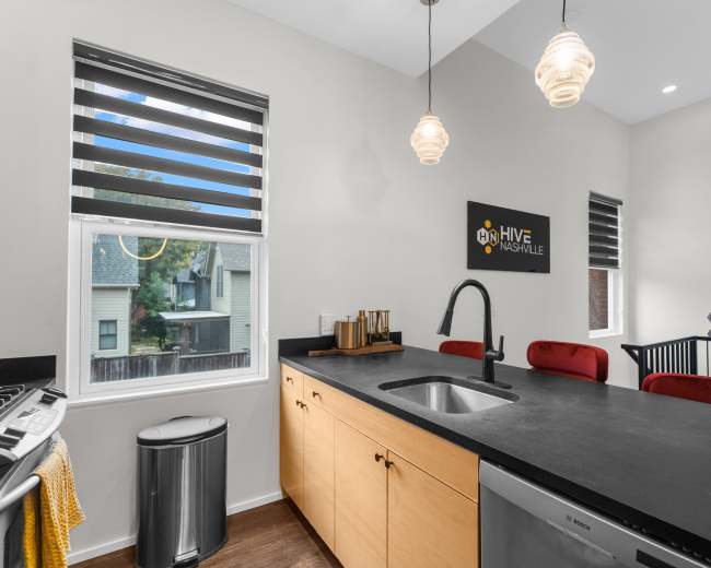 A modern kitchen features a sink and black countertop, with wooden cabinetry and pendant lights, set against a backdrop of large windows and a sign for "Hive Nashville."