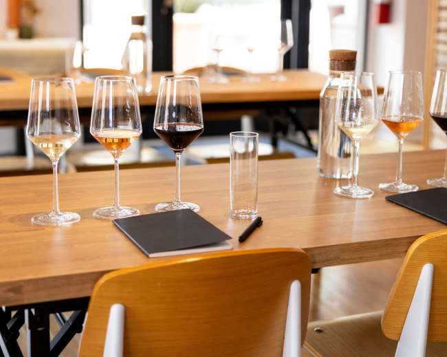 A wooden table is set with several wine glasses and a water bottle, along with notepads and pens for each place setting.