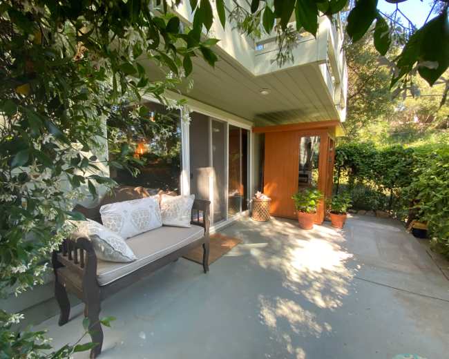 A cozy outdoor seating area with a sofa and potted plants is surrounded by lush greenery and an adjacent wooden entrance.