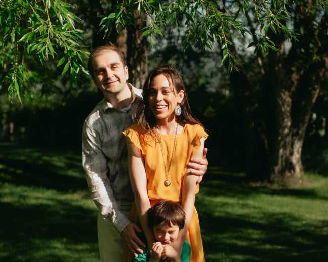 A man and woman stand together with a young boy in front of them, surrounded by trees and grass on a sunny day.