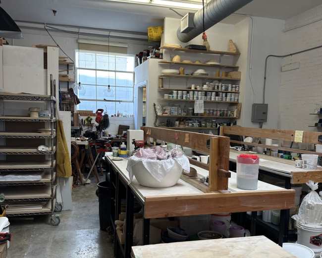 The image shows a pottery studio with various workstations, tools, and materials organized along the walls, all lit by overhead fluorescent lights.