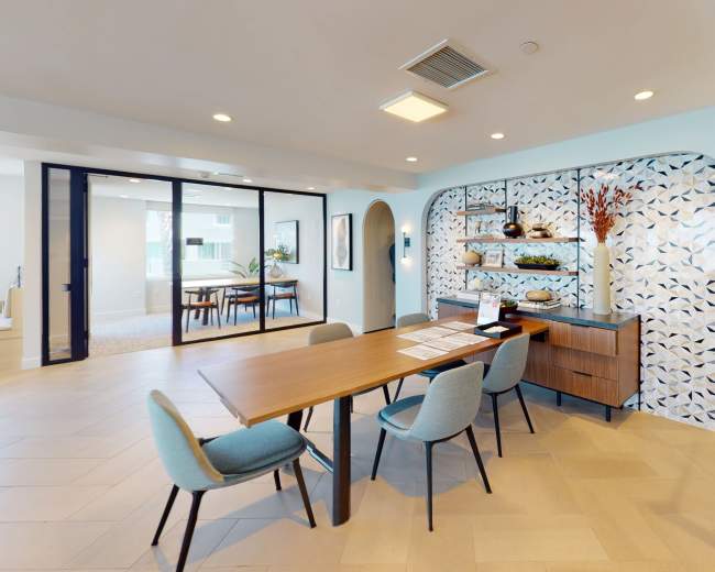 A modern lobby area featuring a central wooden table surrounded by upholstered chairs, with a decorative wall and plants in the background.