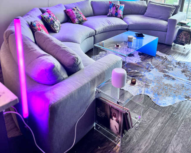 The image shows a modern living room with a gray sectional sofa adorned with colorful pillows, a transparent coffee table, and a neon light feature on the wall.