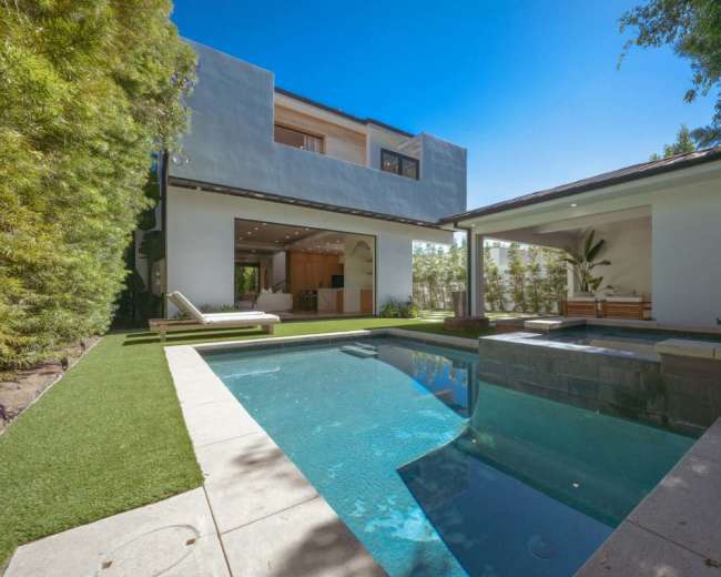 The image shows a modern outdoor space featuring a swimming pool, landscaped greenery, and a house with large windows in the background.